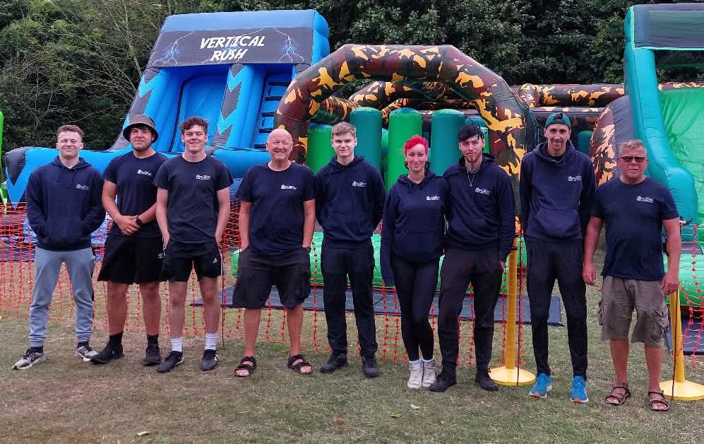 Nine members of The Play and Development Service, all in blue branded clothing, are pictured in front of inflatables in a park. 